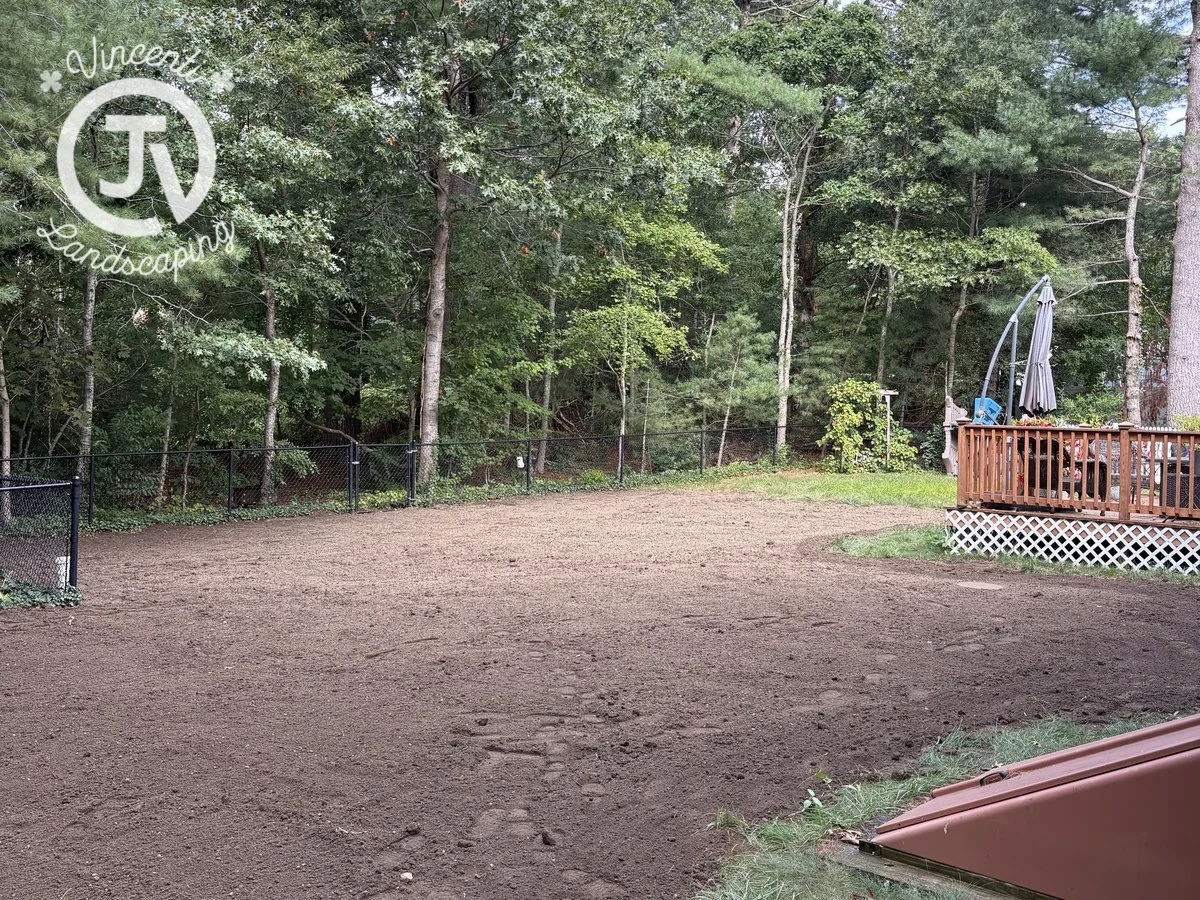 Backyard soil prepared for lawn installation beside a wooden deck and fenced woodland